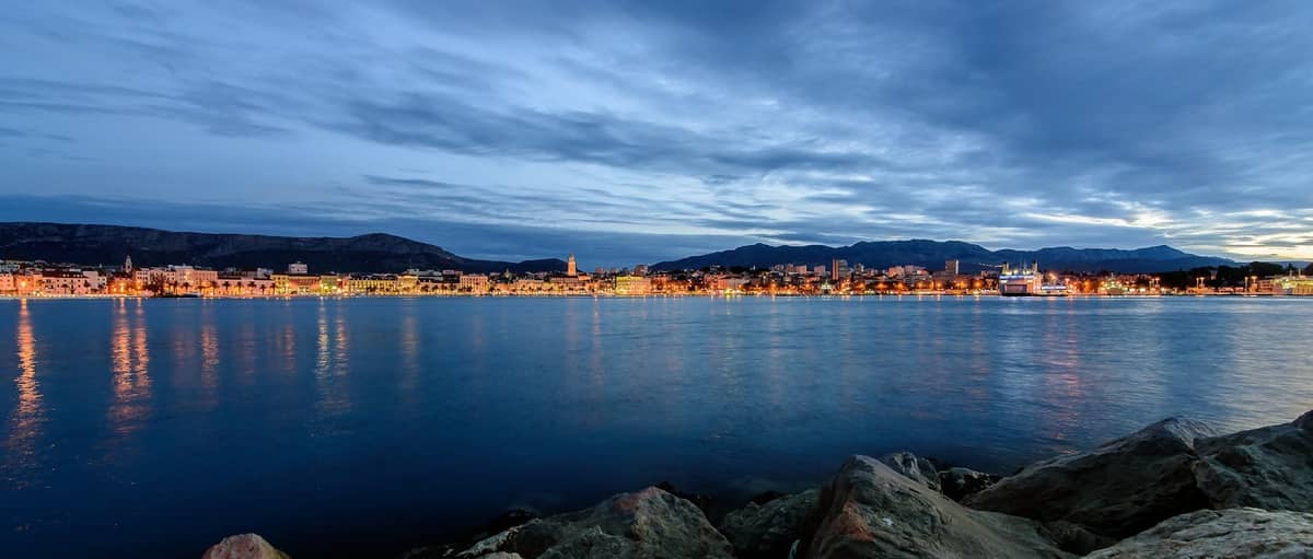 Panoramischer Abendblick auf Splits Uferpromenade mit Stadtlichtern, die sich im Wasser spiegeln, Bergen im Hintergrund und ruhiger Atmosphäre für das Nachtleben.