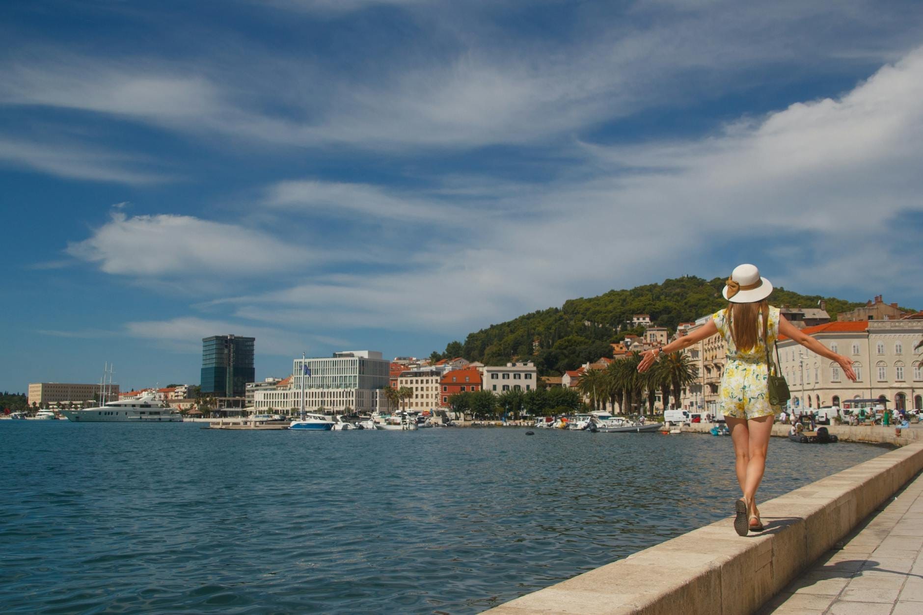 Mulher com vestido de verão caminhando pelo calçadão à beira-mar de Split, Croácia, com barcos, edifícios históricos e a colina Marjan ao fundo.