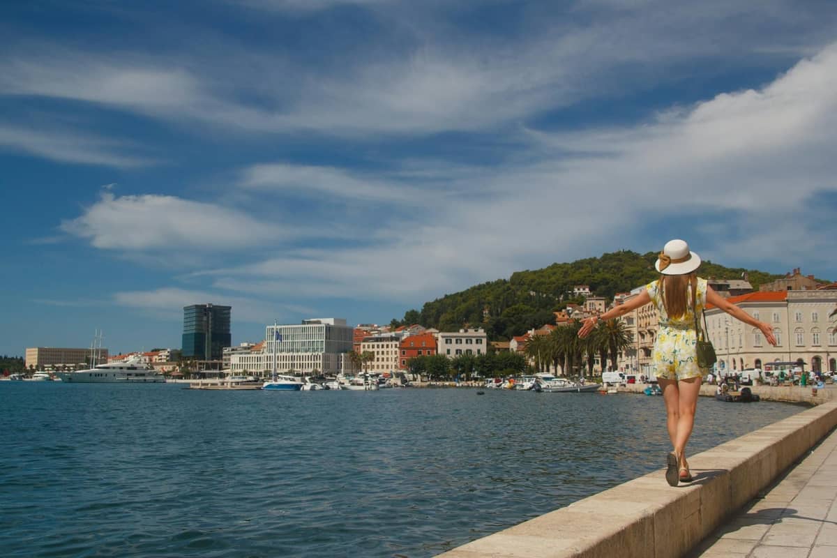 Eine Frau spaziert allein an der Uferpromenade von Split entlang, mit der erkennbaren Stadtsilhouette und dem Yachthafen im Hintergrund unter einem sonnigen Himmel.