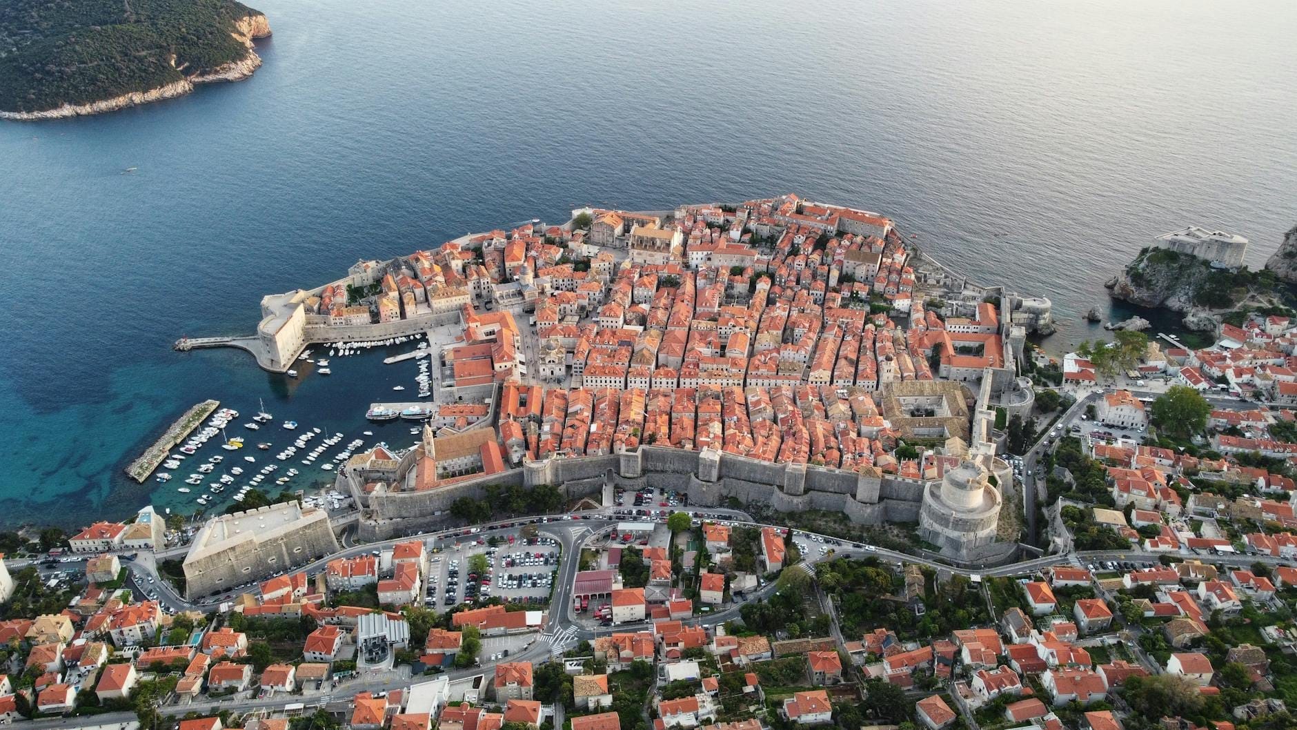 Vista aérea del histórico casco antiguo amurallado de Dubrovnik en la costa del Adriático, con tejados rojos, puerto y murallas de la ciudad visibles frente al mar.