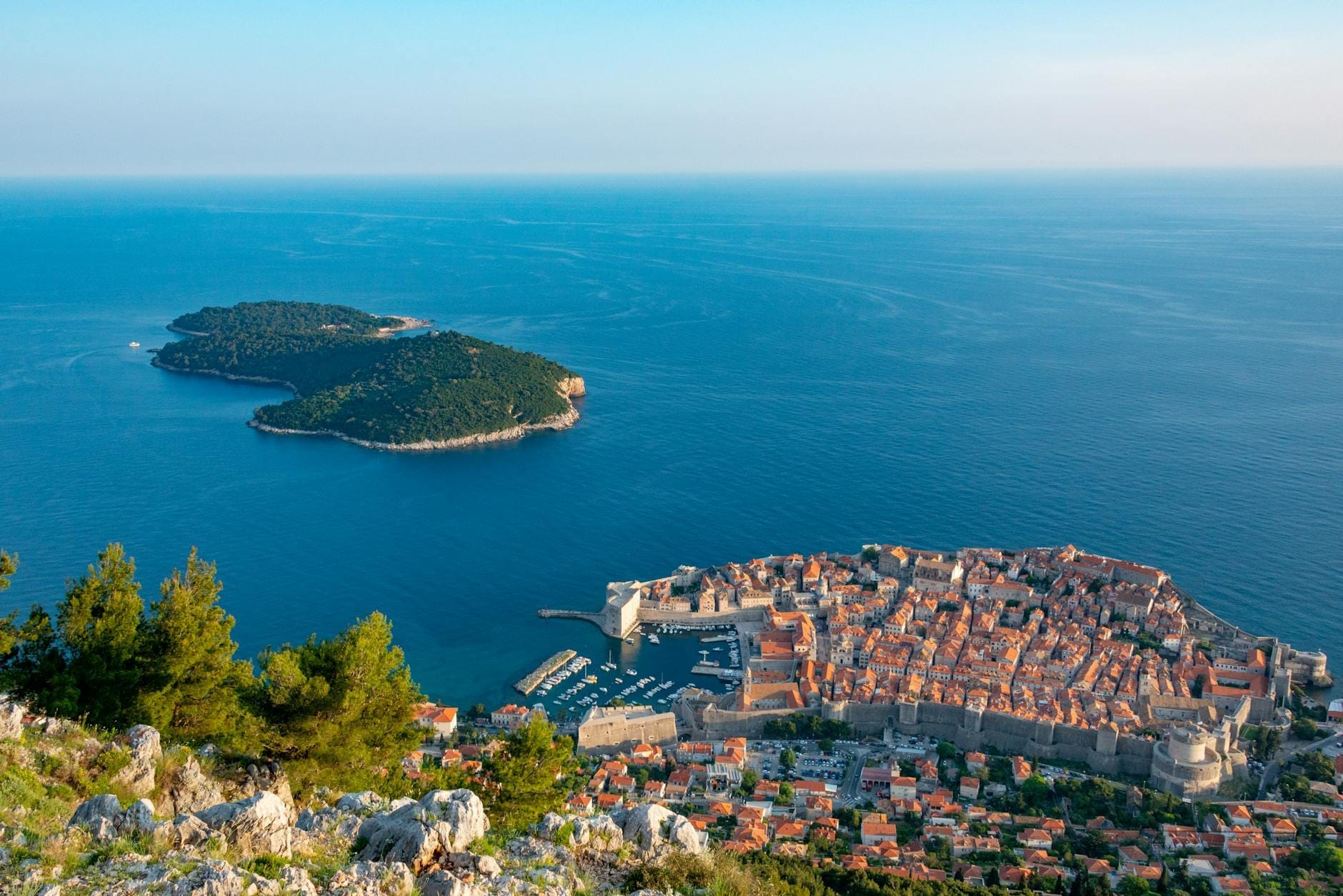 Vista aérea impresionante del casco antiguo de Dubrovnik con tejados naranjas, murallas, mar azul y una isla verde cercana bajo un cielo despejado.