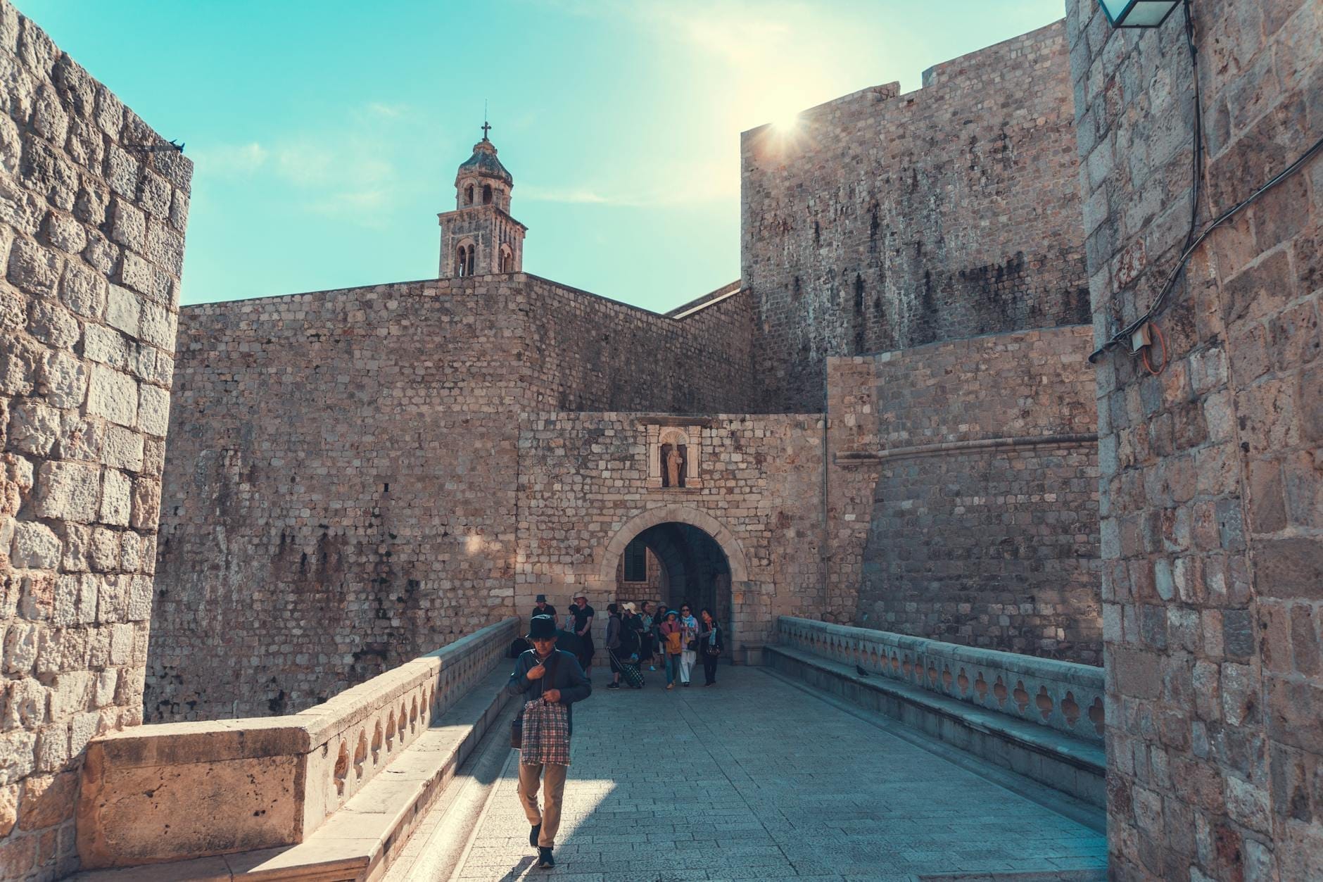 Visitantes caminan sobre un puente de piedra hacia la famosa Puerta Pile, entrada principal al Casco Antiguo de Dubrovnik, bajo un cielo azul despejado.