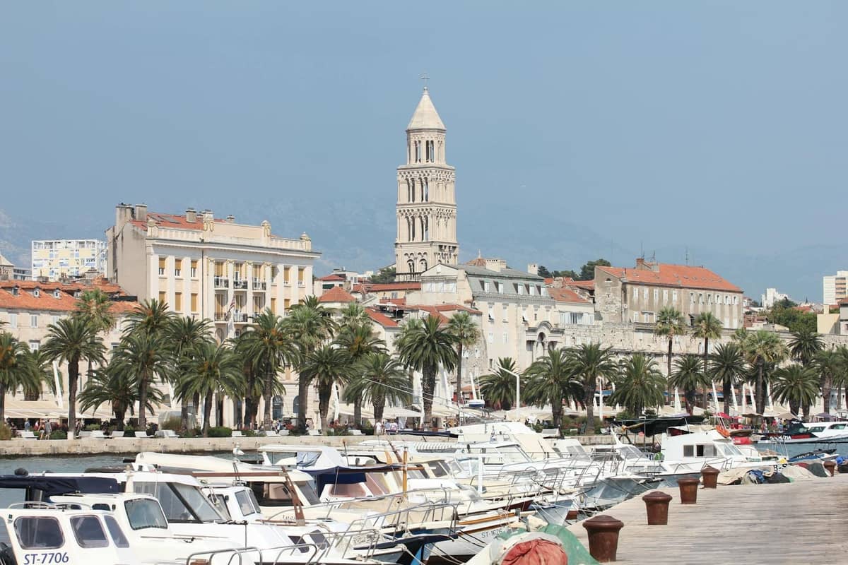 Split waterfront with busy ferry terminal and boats docked in front of palm-lined promenade, Diocletian’s Palace bell tower visible in the background.