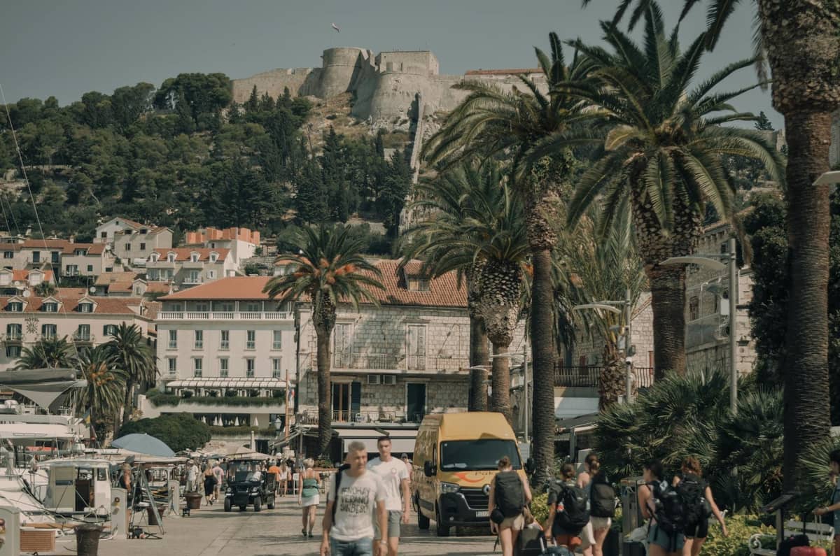 Fußgänger auf der Hafenpromenade von Hvar mit Palmen, historischen Gebäuden und der Festung Hvar auf dem Hügel