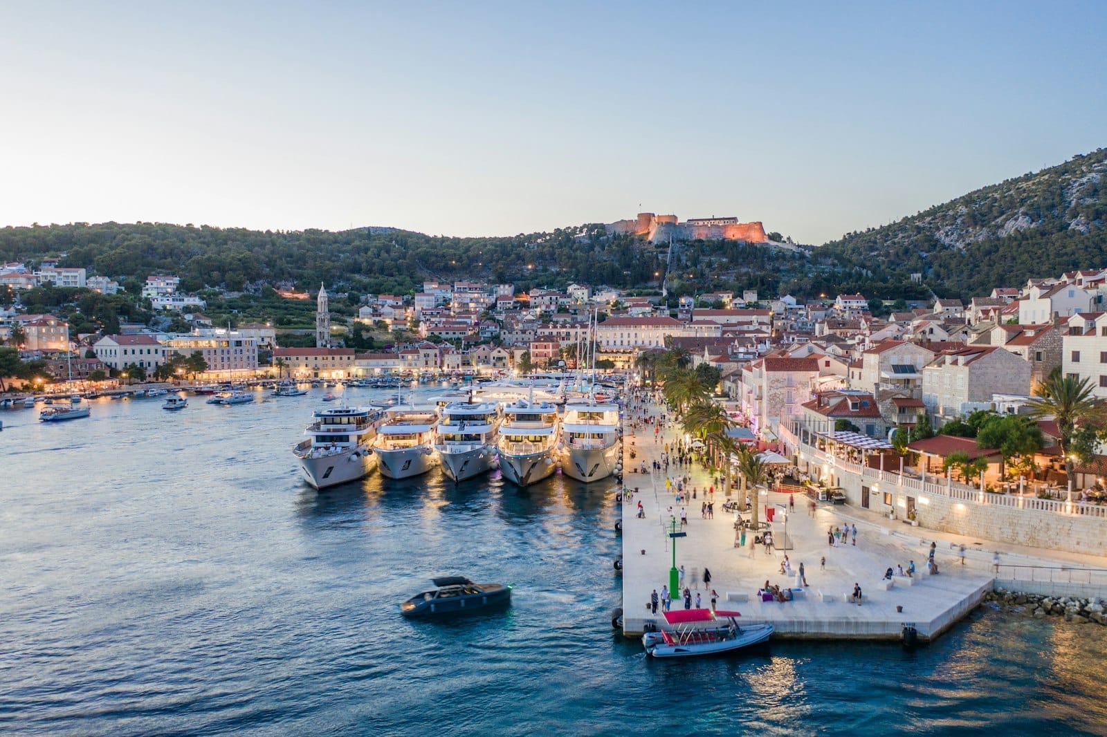 Porto vibrante de Hvar ao pôr do sol com balsas atracadas ao longo da orla, cidade histórica ao fundo e colinas verdes se erguendo atrás.