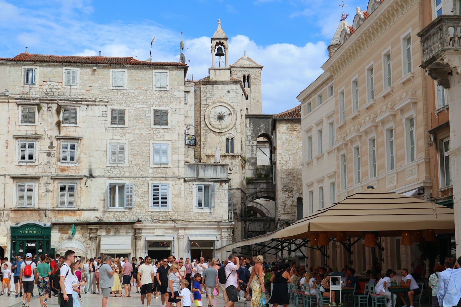 Praça do centro histórico de Split movimentada com turistas, edifícios históricos de pedra, uma torre do relógio e cafés ao ar livre com guarda-sóis num dia ensolarado.