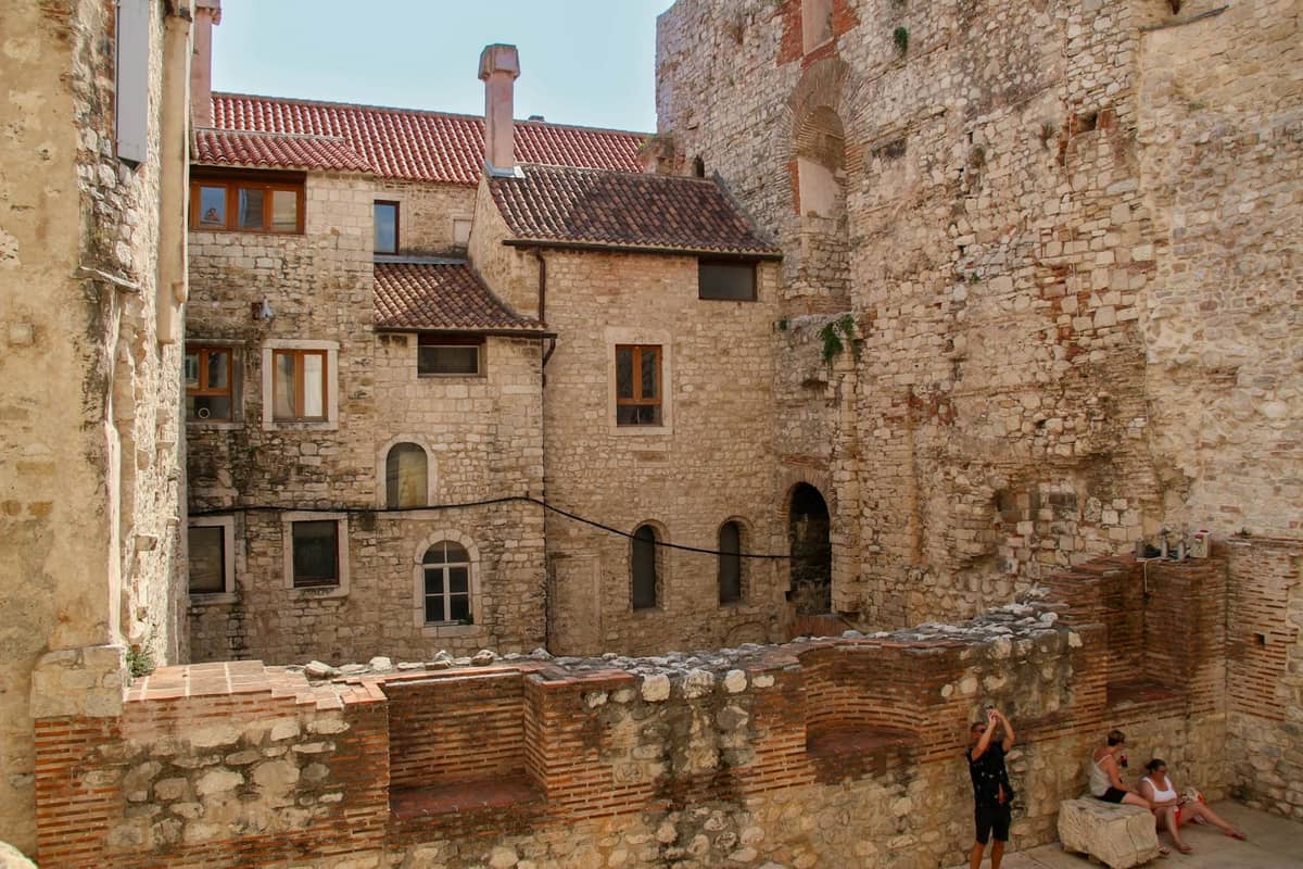 Steinerne Wohngebäude, die in die antiken Palastmauern integriert sind, mit Menschen, die zwischen den Ruinen in der Altstadt von Split sitzen.