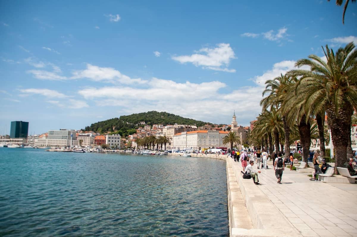 Persone che passeggiano lungo la passeggiata Riva di Spalato, fiancheggiata da palme, acque azzurre, edifici storici e colline in una giornata di sole.