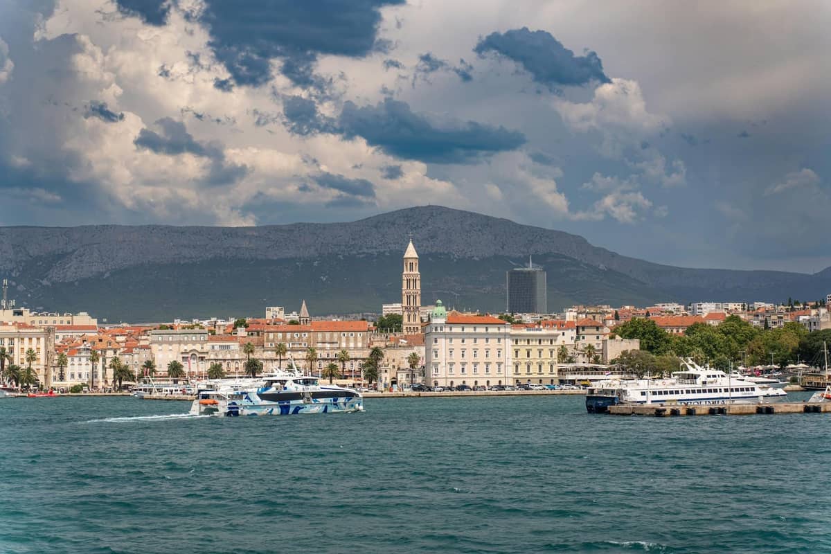 Vista del lungomare storico di Spalato con traghetti e barche sul Mare Adriatico, montagne sullo sfondo e cielo nuvoloso.