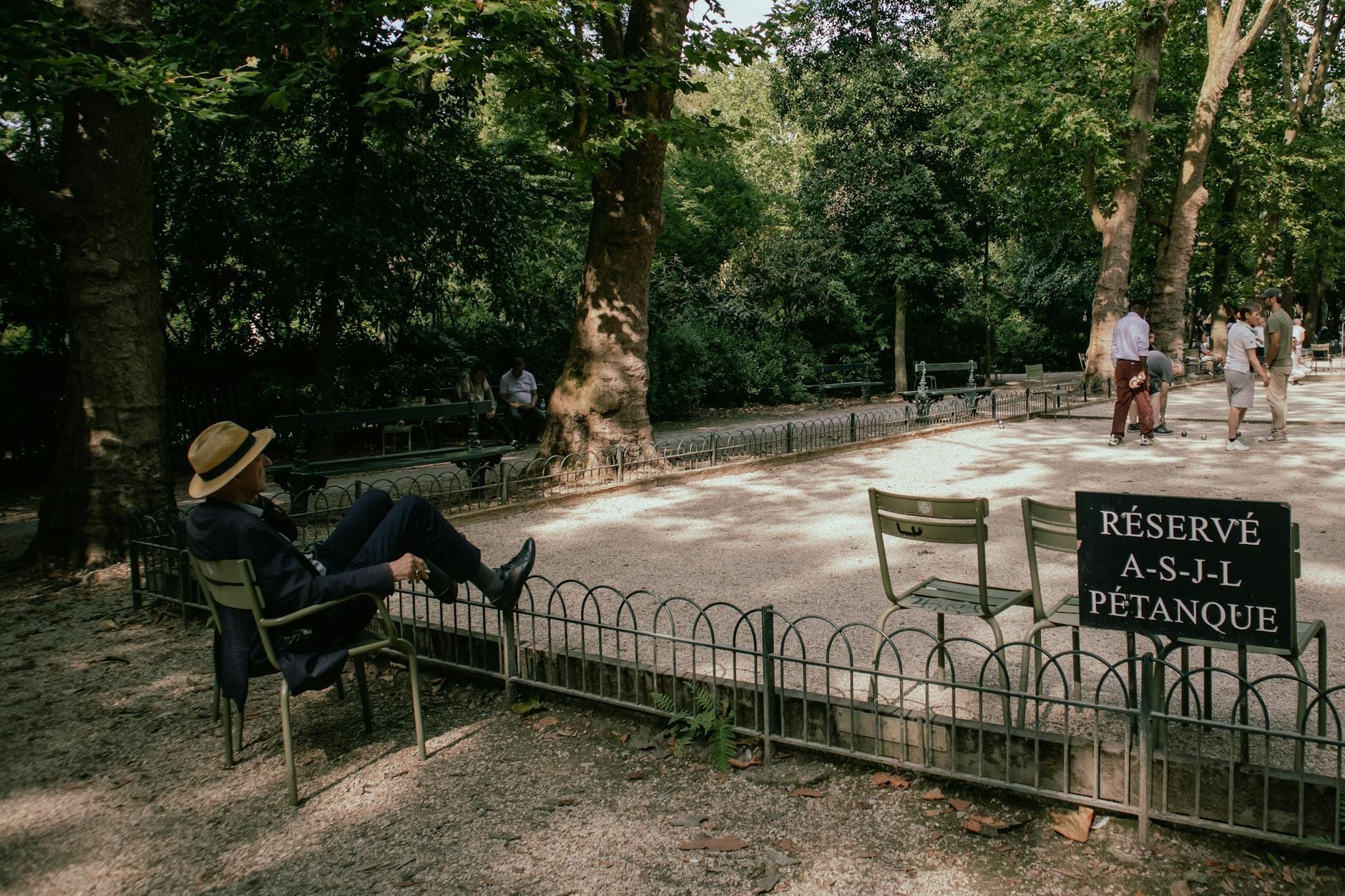 Campo da pétanque all'ombra in un parco verdeggiante, con persone che si rilassano e giocano; una persona con un cappello è seduta su una sedia e un cartello indica 'Réservé pétanque'.