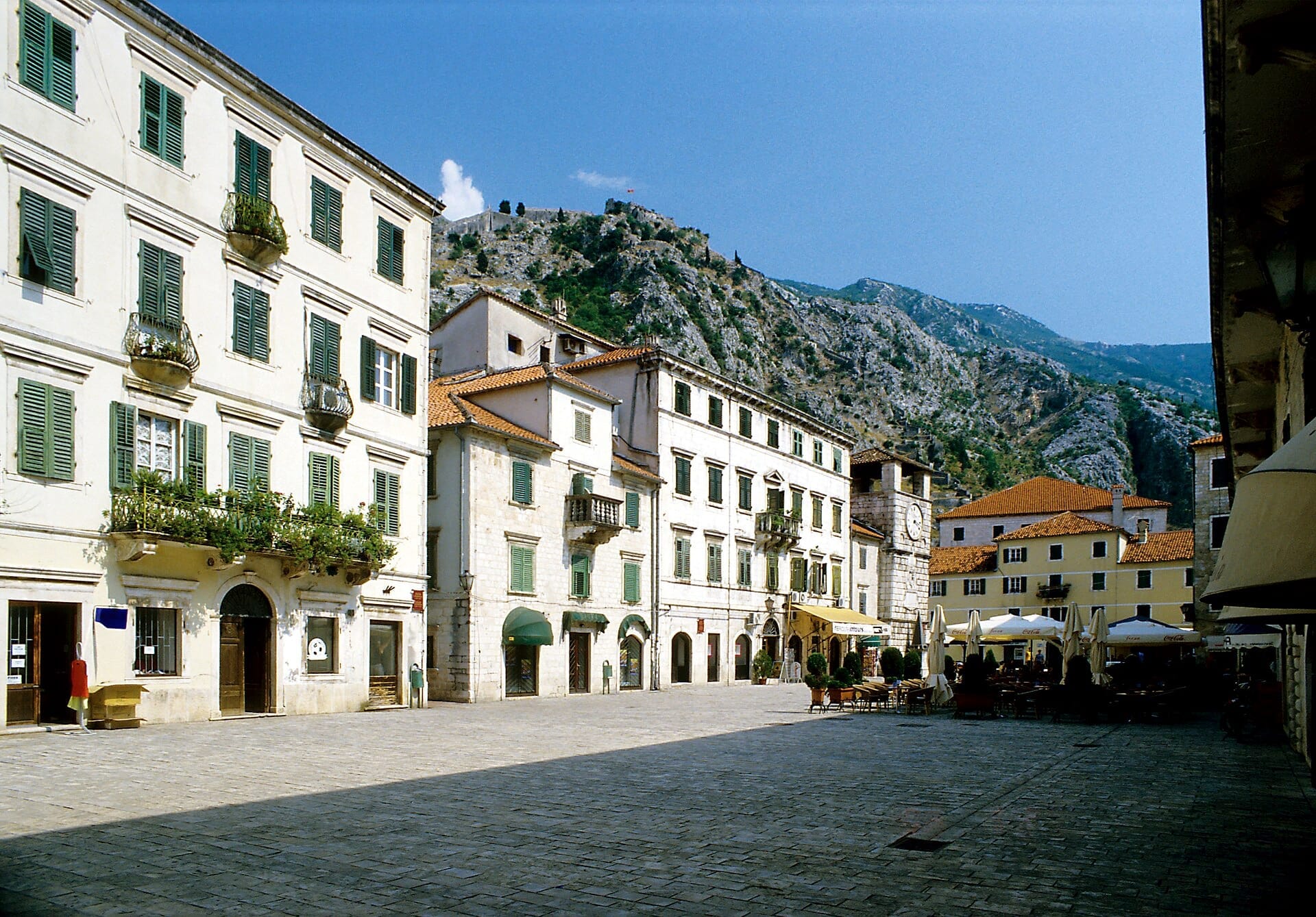 Vista ampia della Piazza delle Armi a Kotor, circondata da storici edifici in pietra con persiane verdi e montagne sullo sfondo.