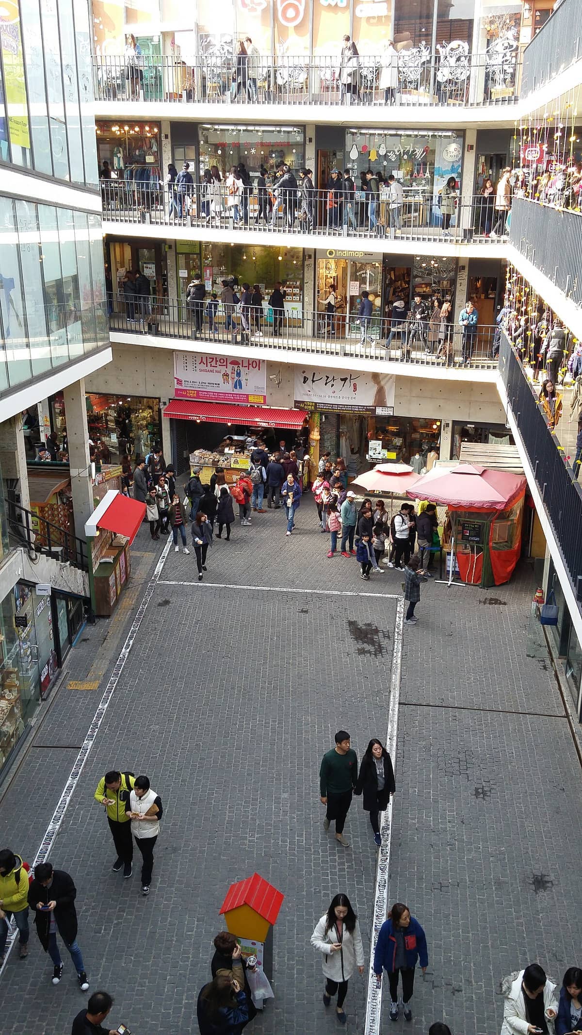 Três andares do pátio aberto de Ssamziegil com pessoas fazendo compras, conversando e caminhando por lojas independentes coloridas e bancas de artesãos durante o dia.