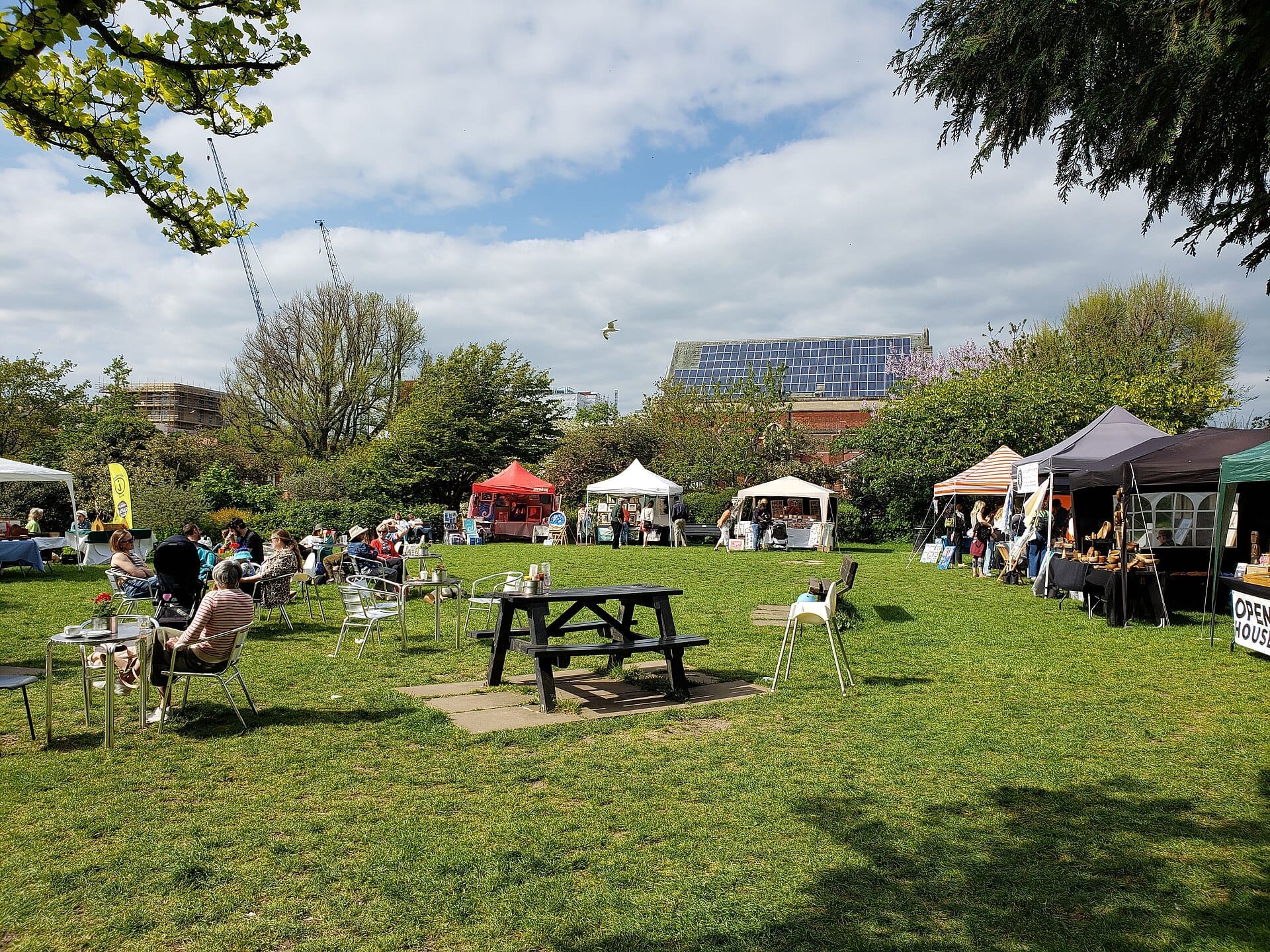 People relax at outdoor tables while local market stalls line the grassy area of St Ann's Well Gardens under a partly cloudy sky.