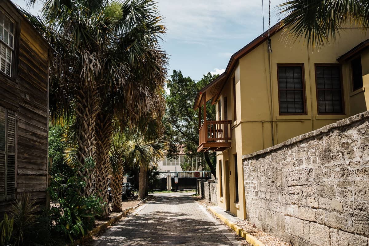 Enge Kopfsteinpflasterstraße im St. Augustine Historic District mit Palmen, historischer Steinmauer und einem alten gelben Gebäude im Mittagslicht.