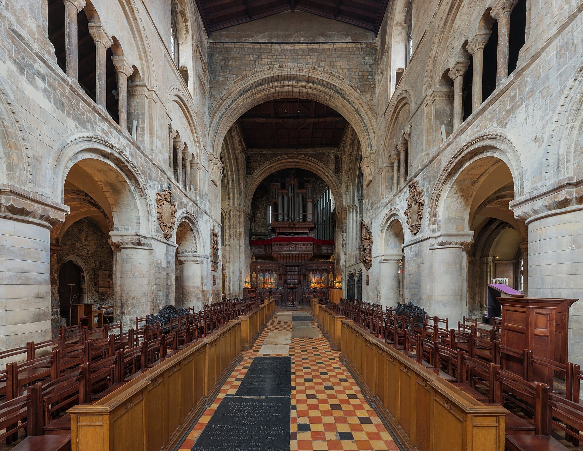L'interno della chiesa St Bartholomew the Great a Londra, con archi normanni, colonne in pietra, banchi in legno e un pavimento di piastrelle decorate.