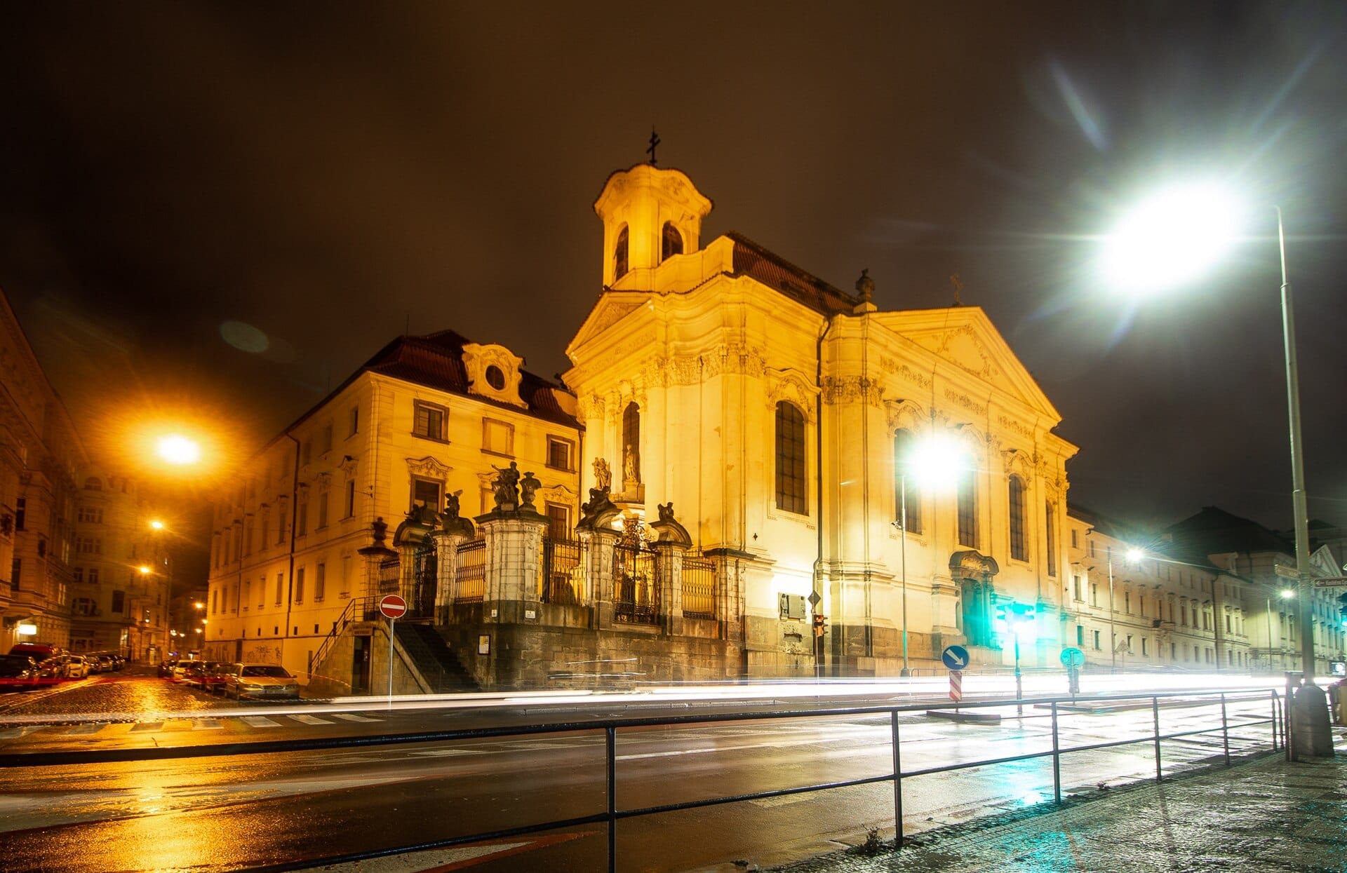 Die Kathedrale der Heiligen Kyrill und Method in Prag bei Nacht beleuchtet, mit leuchtenden Straßenlaternen und nassen Spiegelungen auf der umliegenden Straße.