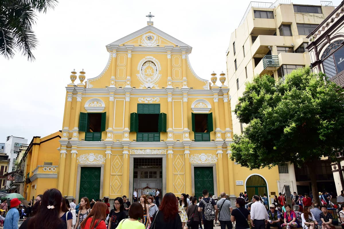 Die gelbe Barockfassade der São-Domingos-Kirche in Macau mit grünen Fensterläden und weißen Zierleisten, davor eine Menschenmenge.