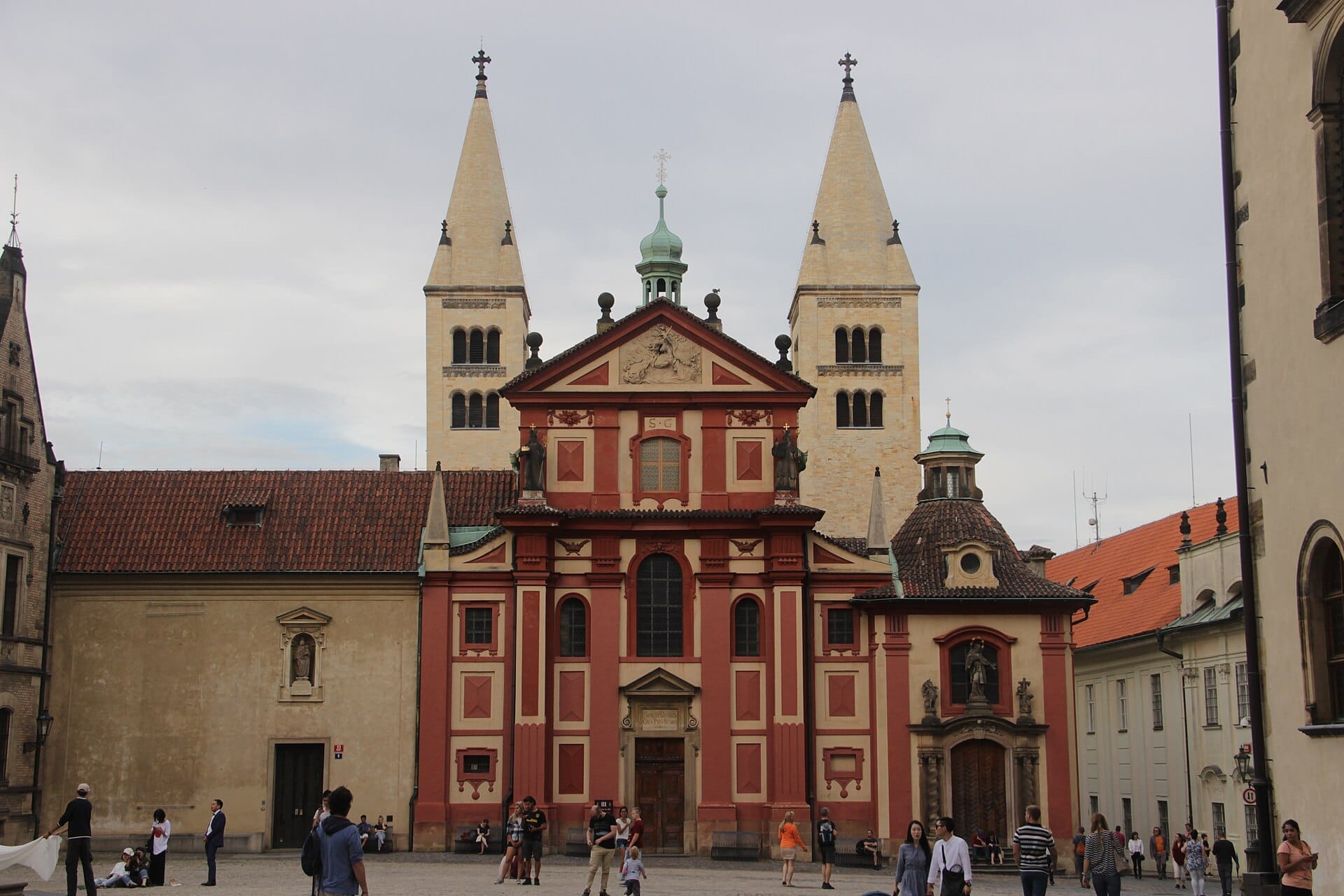La Basilica di San Giorgio al Castello di Praga con la sua caratteristica facciata rossa, le torri gemelle romaniche e i visitatori che passeggiano nel cortile sotto un cielo nuvoloso.