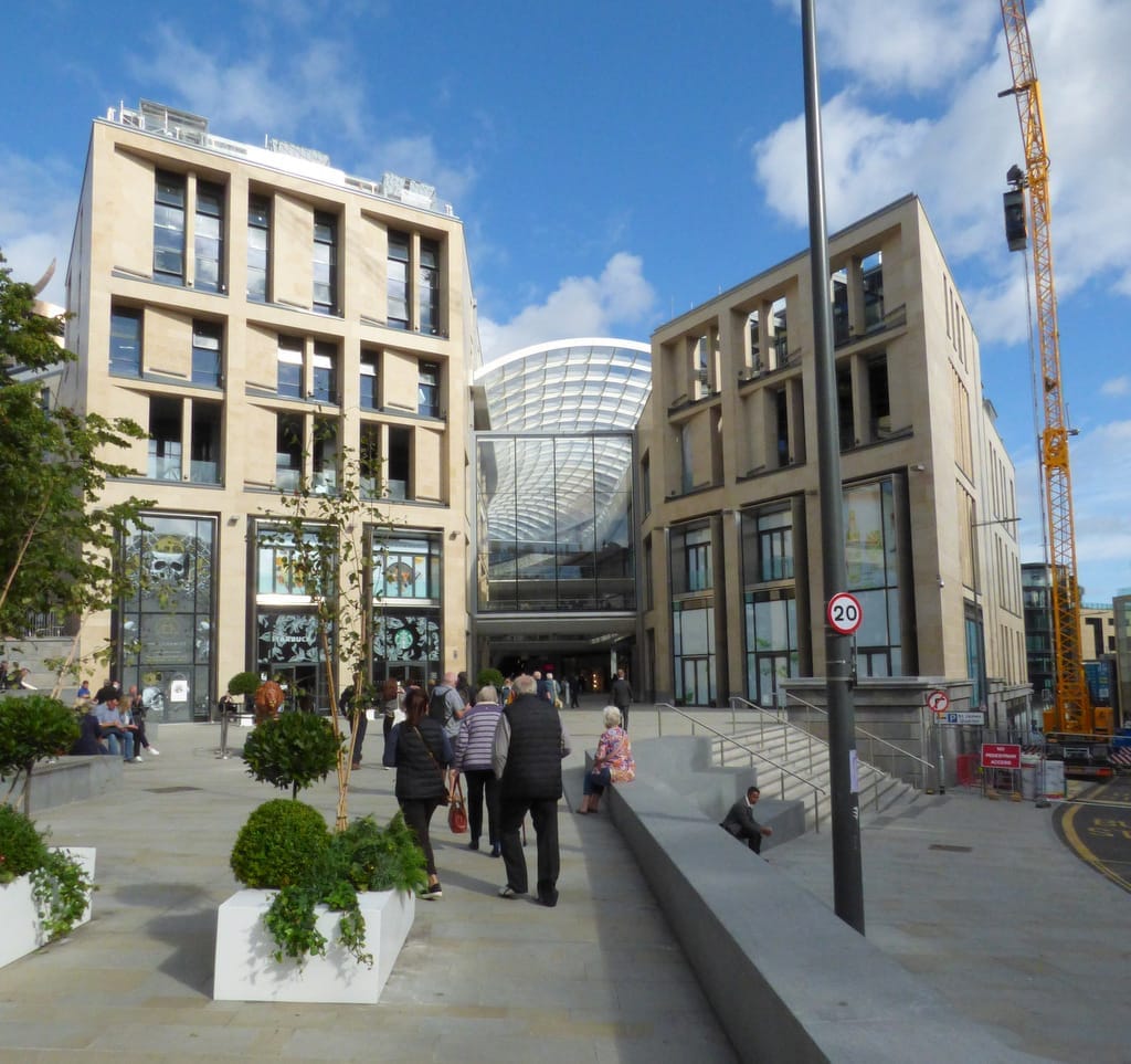 Moderne Sandsteingebäude des St James Quarter mit einem geschwungenen Glasdach, Menschen, die an einem hellen, klaren Tag entlang einer breiten Fußgängerzone spazieren.