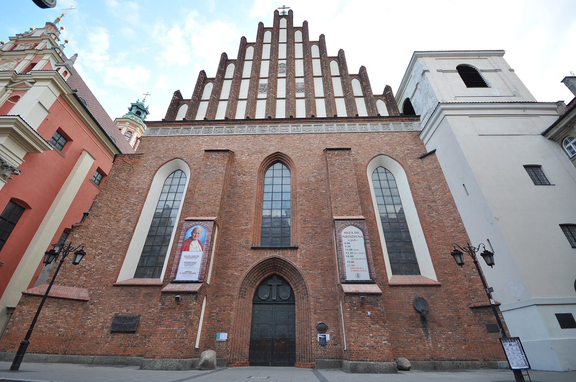 มุมมองด้านหน้าของมหาวิหาร St. John's Archcathedral ในกรุงวอร์ซอ โดดเด่นด้วยหน้าอาคารอิฐสูง หน้าต่างสไตล์โกธิก และป้ายแบนเนอร์ทั้งสองข้างทางเข้า