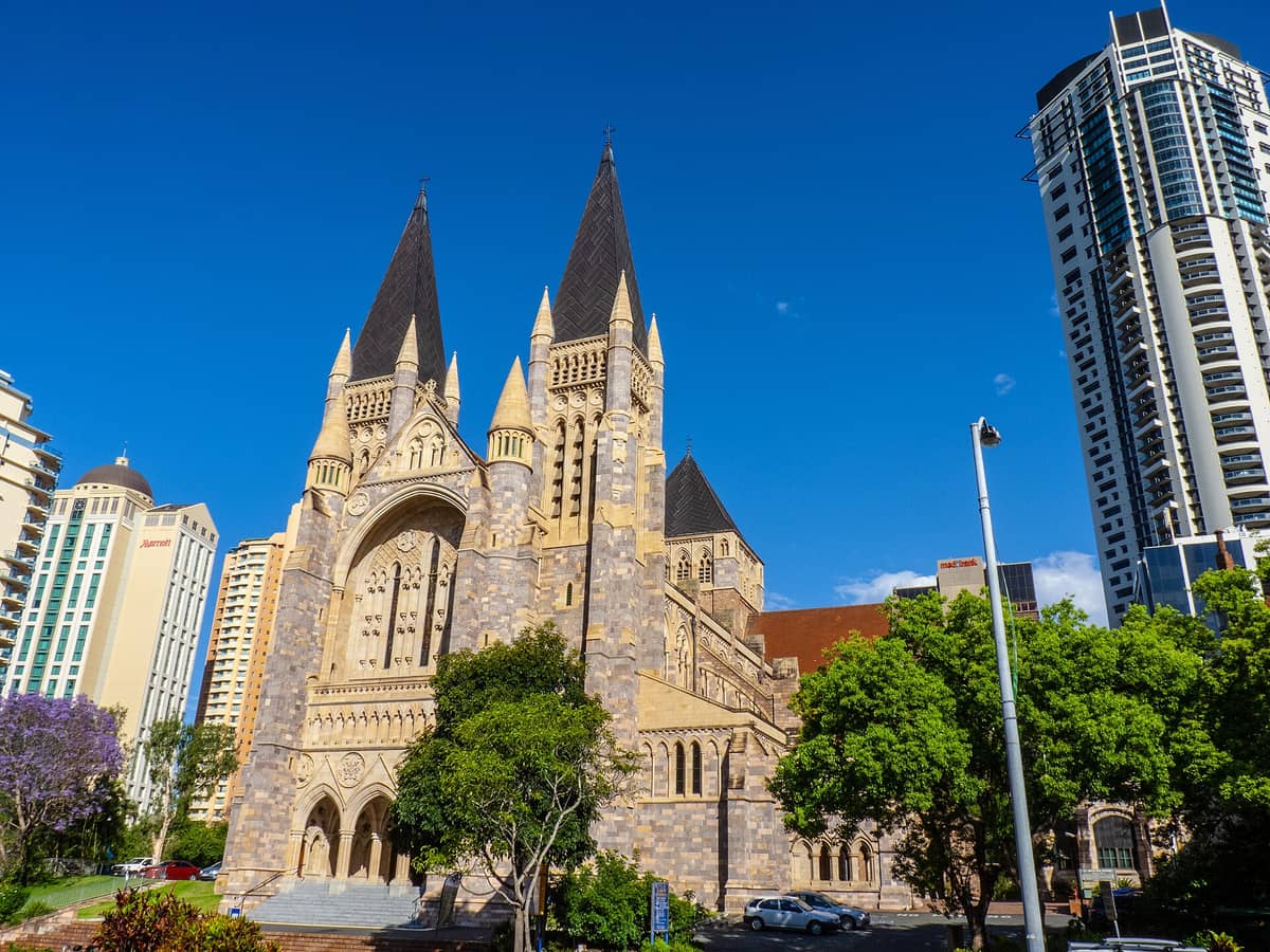 St John's Cathedral in Brisbane mit ihren markanten neugotischen Zwillingstürmen aus Stein, umgeben von modernen Stadtgebäuden und Bäumen unter strahlend blauem Himmel.