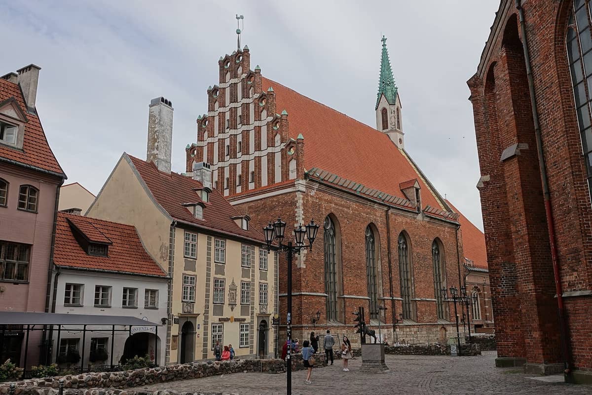 Außenansicht der Johanniskirche in Riga mit ihrer mittelalterlichen Backsteinfassade, dem Treppengiebel und dem hohen Turm mit grüner Turmspitze, umgeben von Altstadtgebäuden.