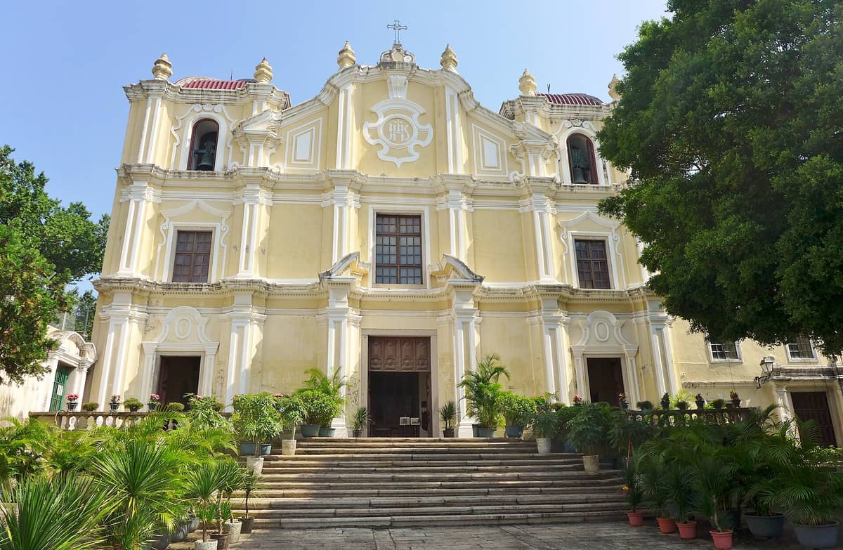 Frontansicht des Priesterseminars und der Kirche St. Joseph in Macau mit der gelben Barockfassade, der Freitreppe und üppigen Topfpflanzen unter einem strahlend blauen Himmel.