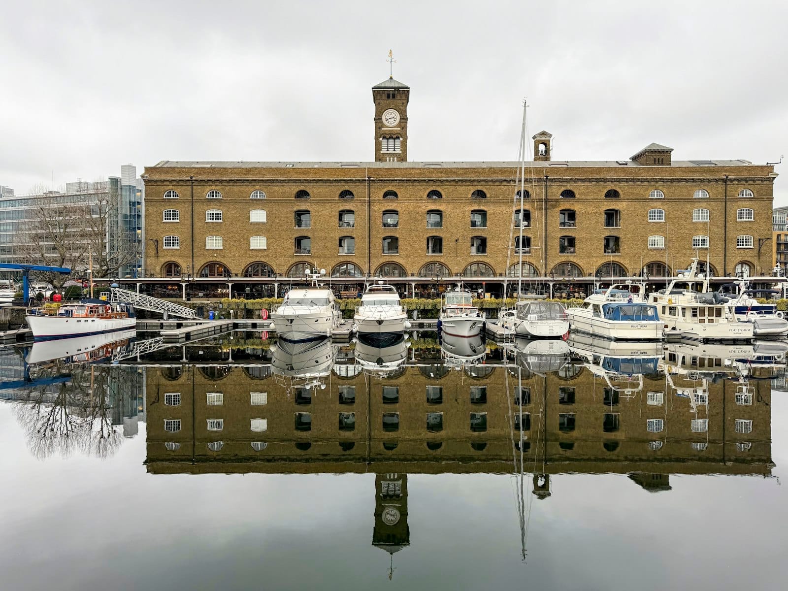 Marina di St Katharine Docks con storico magazzino in mattoni, torre dell'orologio e barche ormeggiate riflesse nell'acqua calma in una giornata nuvolosa.