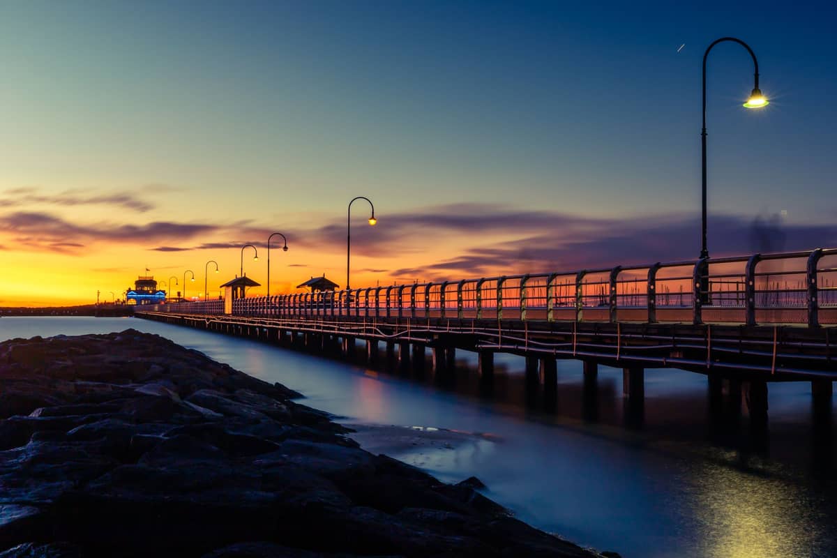 Der St Kilda Pier bei Sonnenuntergang: ein langer Steg erstreckt sich über das Wasser, gesäumt von Laternen und fernen Pavillons unter einem farbenprächtigen Himmel.