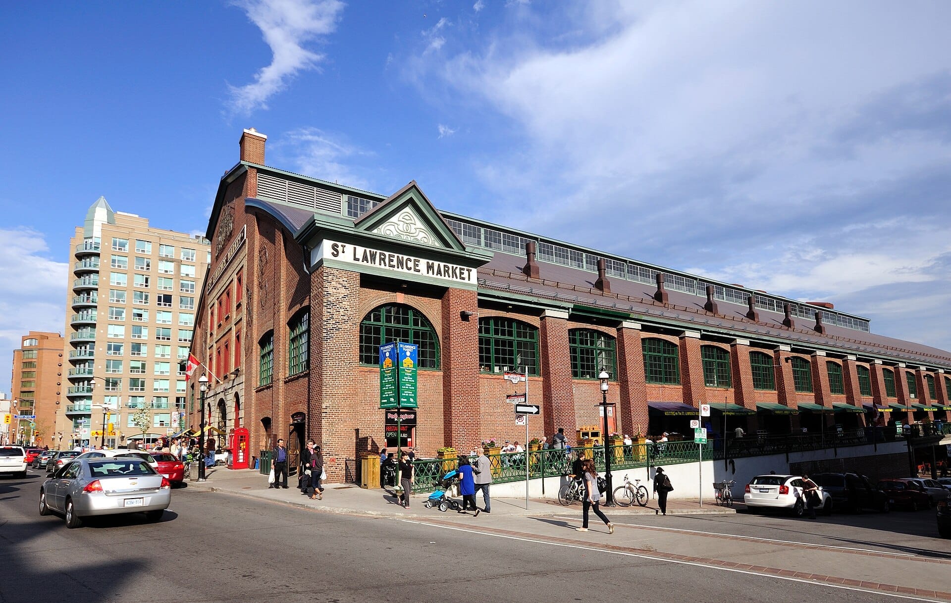 อาคาร St. Lawrence Market ในโตรอนโต มีผนังภายนอกอิฐสีแดง หน้าต่างสีเขียวขนาดใหญ่ และผู้คนกำลังเดินในวันที่แดดสดใส
