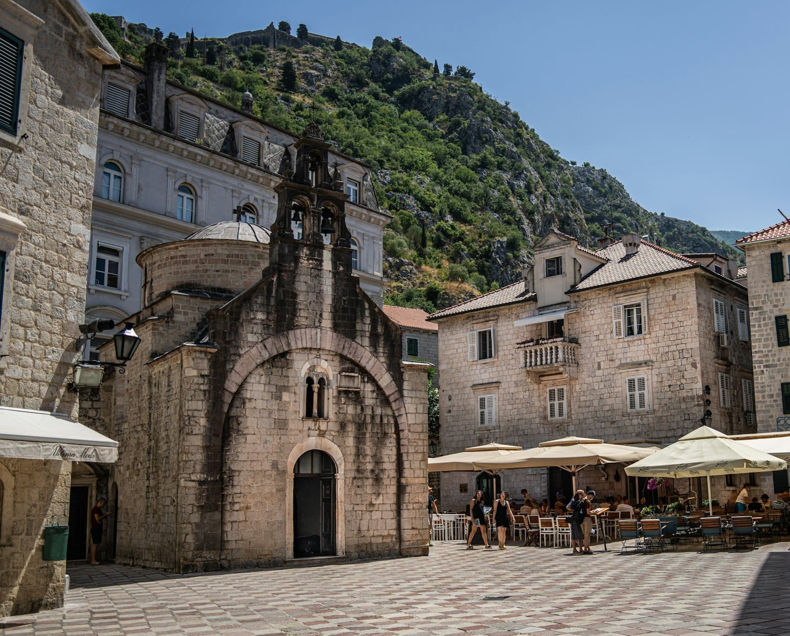 La Chiesa di San Luca nella Città Vecchia di Kotor, circondata da edifici in pietra, caffè all'aperto e colline verdi sotto un cielo azzurro e sereno.