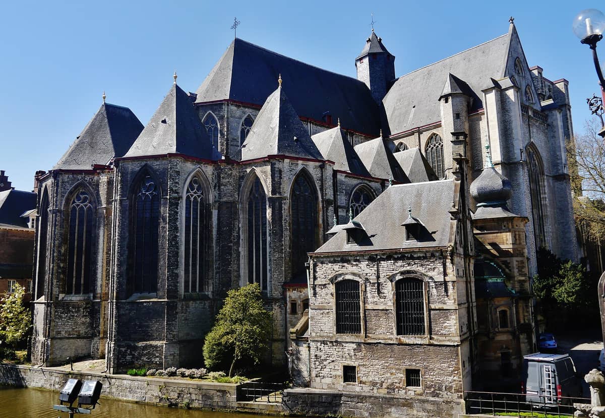 St Michael's Church in Ghent, with its distinctive unfinished tower and detailed Gothic stonework, viewed from across the Leie canal on a clear day.