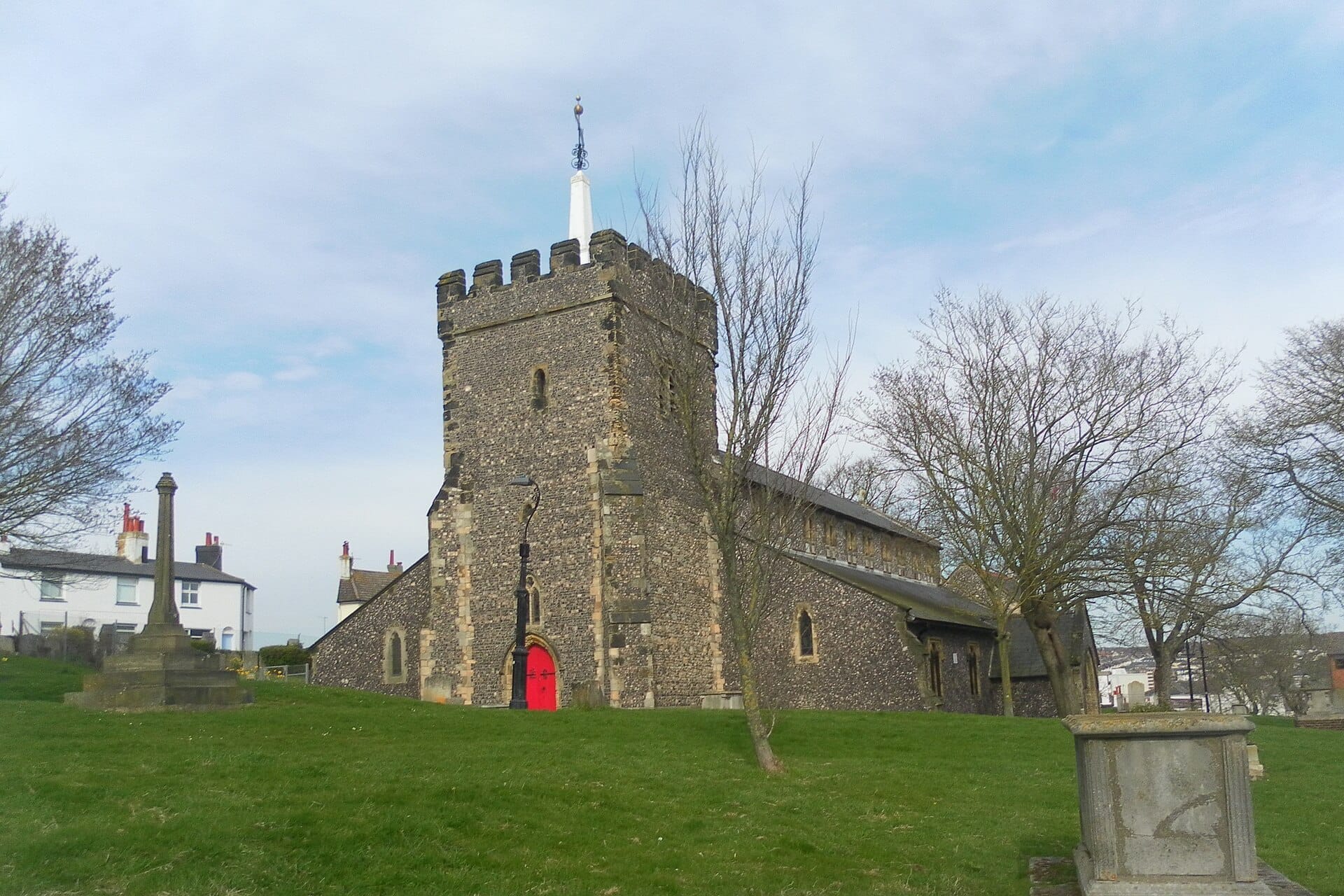 St Nicholas of Myra church in Brighton, an ancient stone building with a battlemented tower and red doors, surrounded by grassy churchyard and leafless trees.