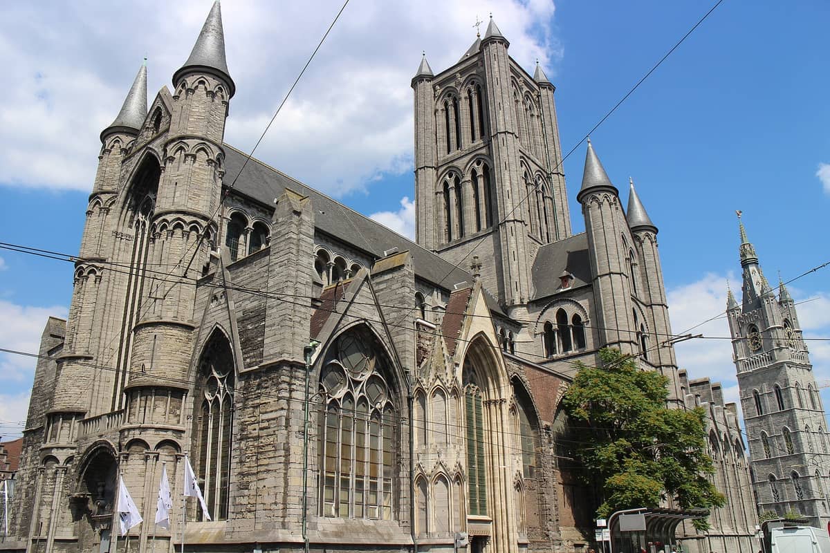 St Nicholas' Church in Ghent with its signature Gothic spires and stone facade under a blue sky, tram wires crossing in front.
