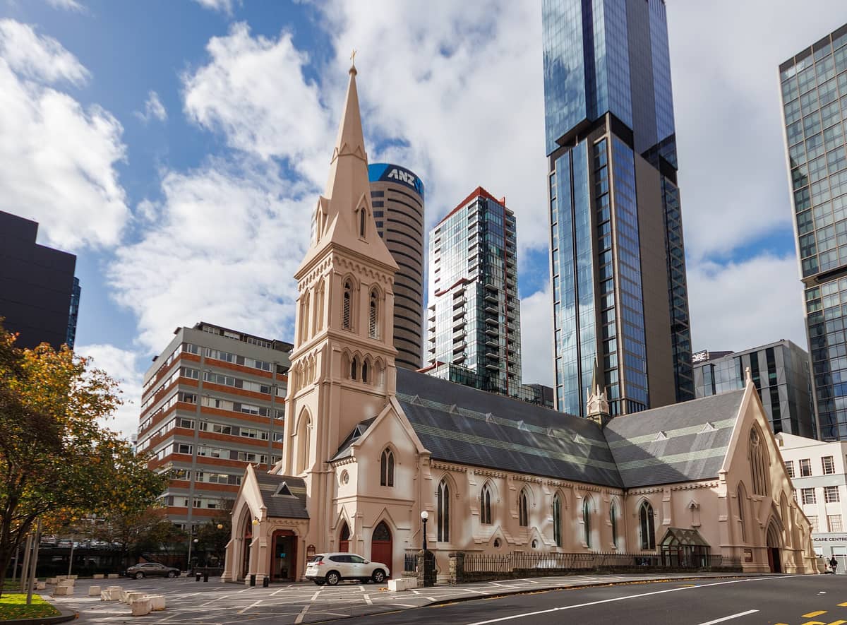 St Patrick's Cathedral in Auckland, eine cremefarbene neugotische Kirche mit einem hohen Turm, umgeben von modernen Stadtgebäuden unter einem teils bewölkten Himmel.