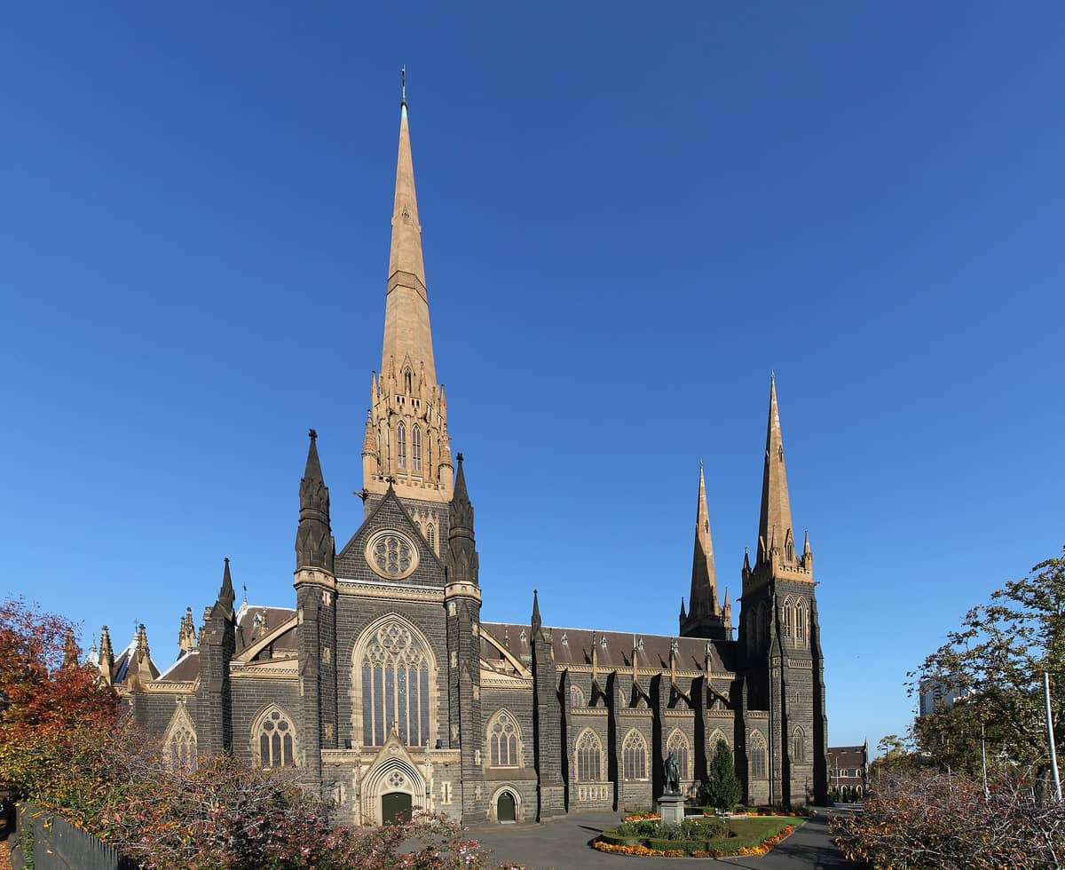 Vista amplia del exterior de la Catedral de San Patricio de Melbourne, con su alta aguja central y su arquitectura neogótica contra un cielo azul despejado.