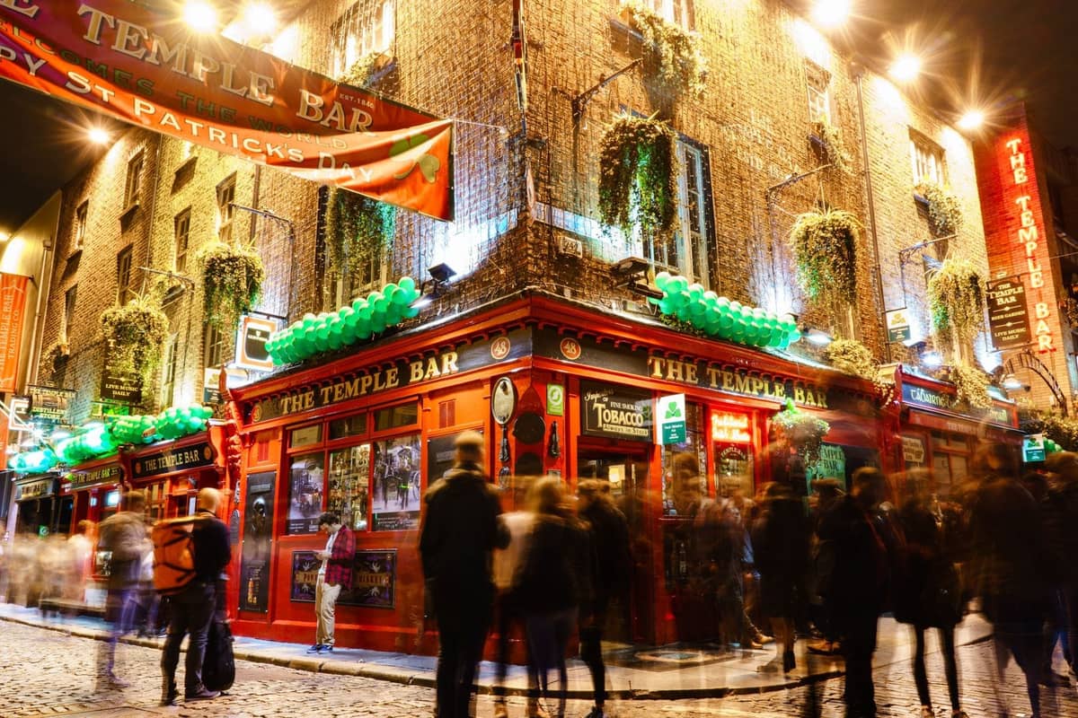 Vista notturna del Temple Bar a Dublino con striscioni del Giorno di San Patrizio, decorazioni verdi e folla radunata all'esterno.