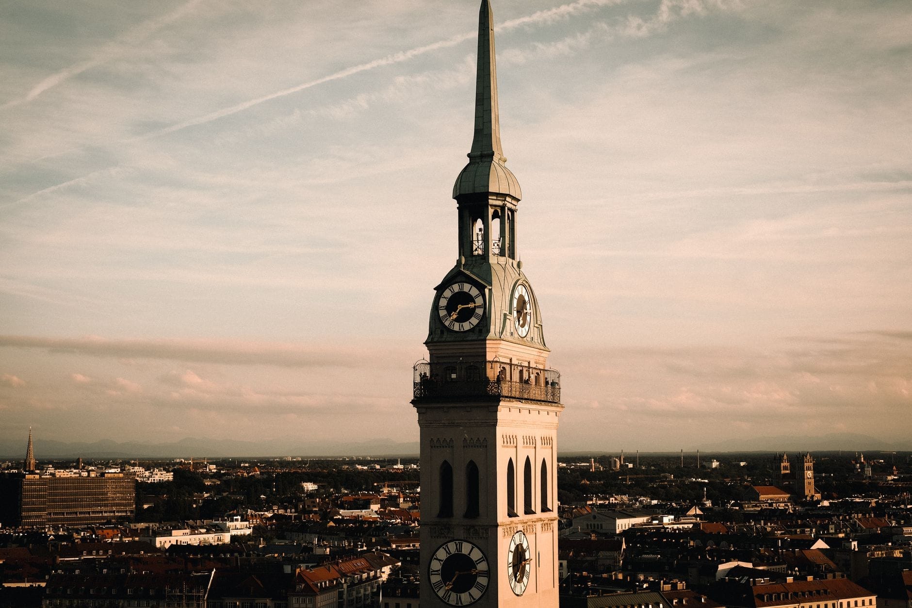 หอคอยโบสถ์ St. Peter's Church สูงตระหง่านเหนือเส้นขอบฟ้าเมือง München ยามพระอาทิตย์ตกดิน พร้อมหน้าปัดนาฬิกาและเทือกเขาที่อยู่ไกลออกไปใต้ท้องฟ้าสีพาสเทล