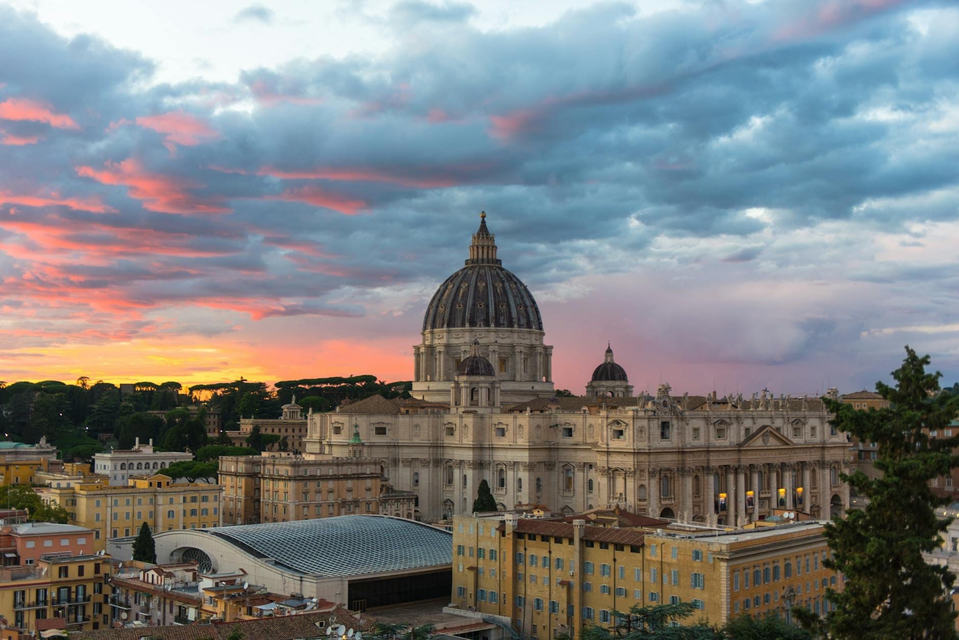 A dramatic sunset view of St. Peter's Basilica rising above the cityscape of Rome, with colorful clouds and rooftops in the foreground.