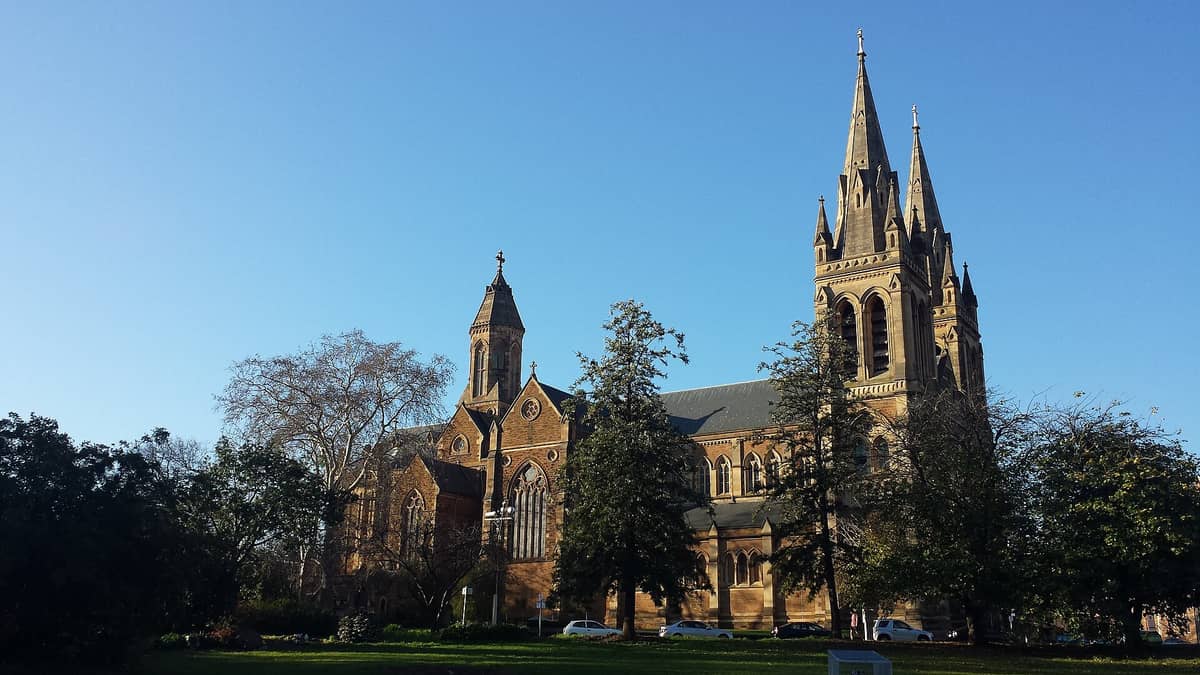 Wide view of St Peter's Cathedral in North Adelaide, showing its twin Gothic spires, large stained glass windows and surrounding greenery in clear daylight.