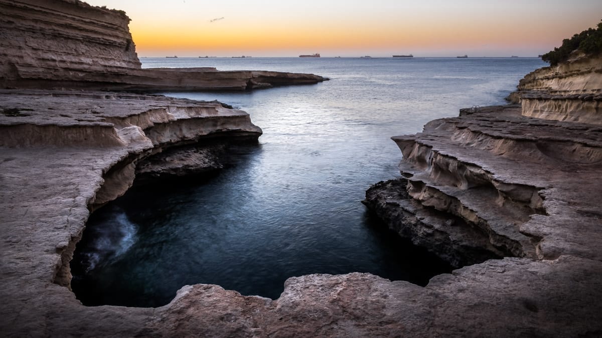 St. Peter's Pool, una baia naturale a ferro di cavallo in pietra calcarea sulla costa di Malta, con sporgenze rocciose stratificate e mare calmo al tramonto.