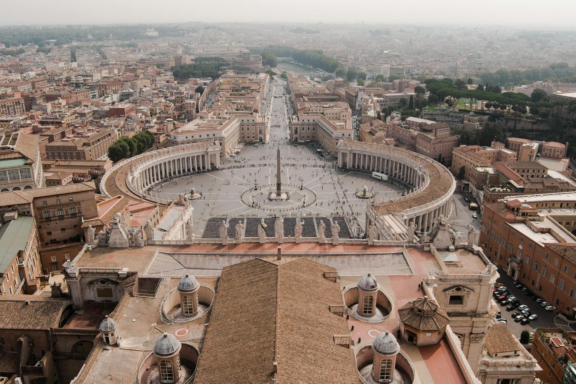 A sweeping aerial view of St. Peter's Square and the surrounding city of Rome, showing the iconic elliptical colonnades and central obelisk bathed in natural daylight.