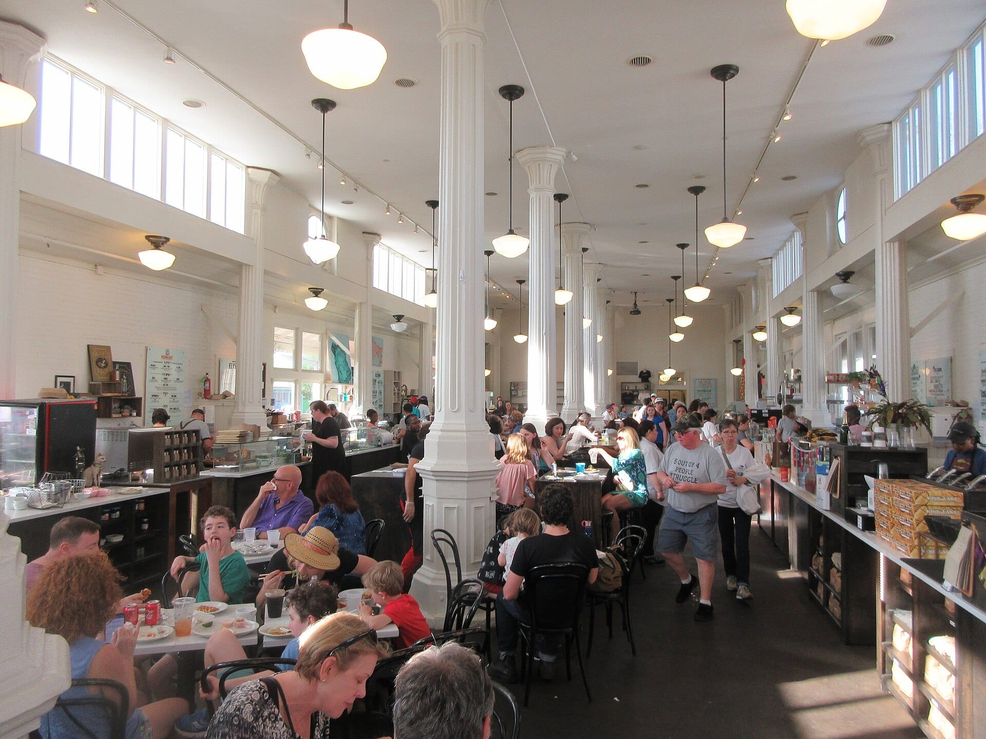 Interno affollato del St. Roch Market con luce naturale, alte colonne bianche, clienti ai tavoli comuni e venditori dietro i banconi a New Orleans.