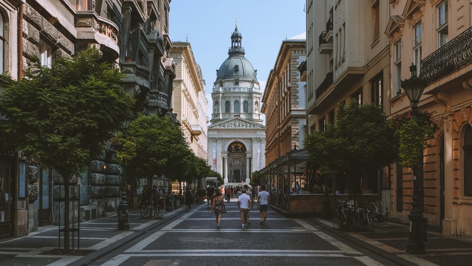 Ampia strada pedonale che conduce direttamente alla Basilica di Santo Stefano a Budapest, con persone che camminano e alberi ai lati sotto un cielo azzurro e limpido.