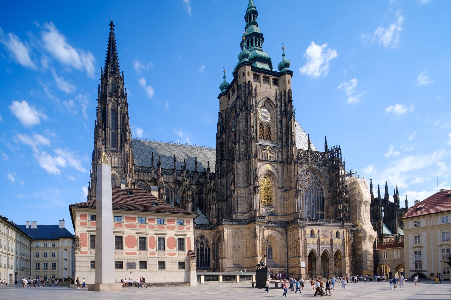Weitwinkelblick auf die prächtige gotische Außenfassade des Veitsdoms mit Türmen und Uhrturm vor blauem Himmel, umgeben von Besuchern auf dem Gelände der Prager Burg.
