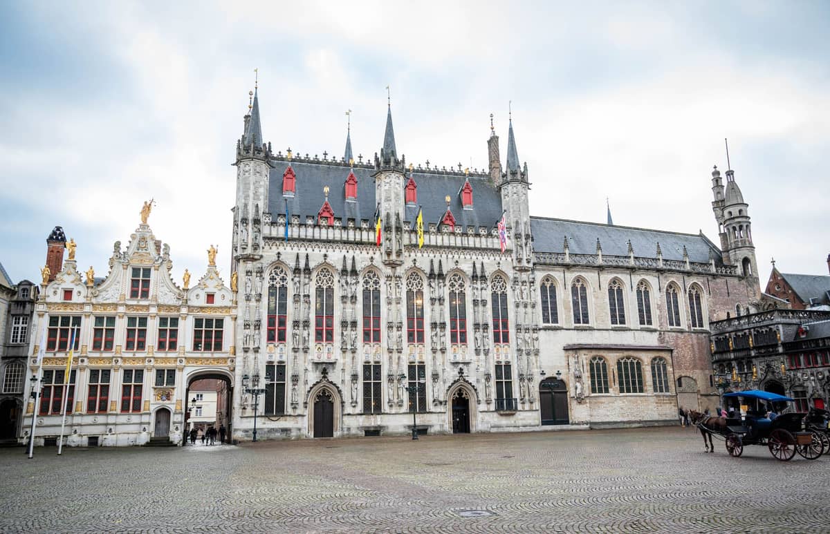 Wide-angle view of the elaborate Gothic facade of Bruges City Hall on Burg Square, showing intricate details and spires, with the adjacent buildings and cobblestone plaza in front.