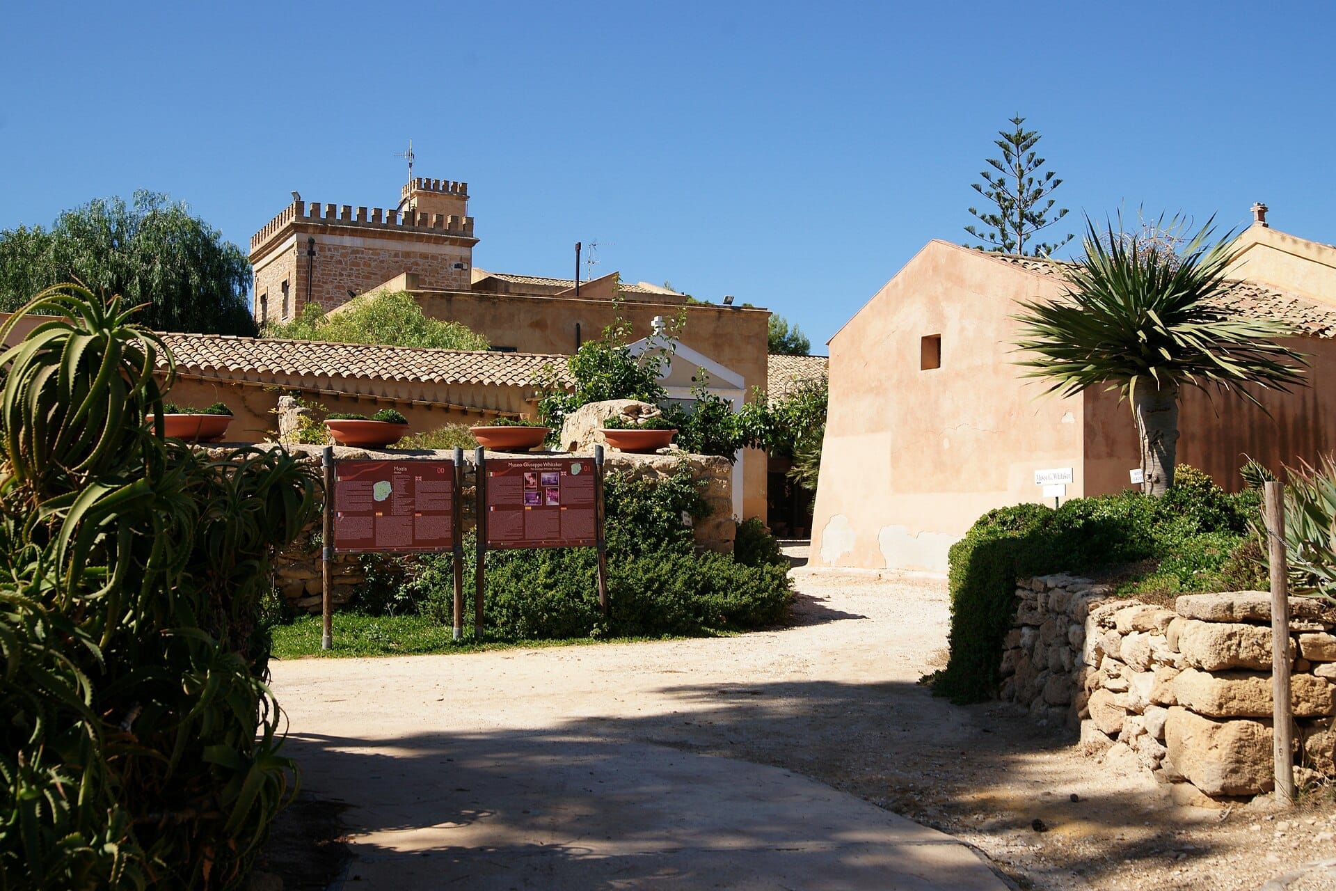 Historische Gebäude und Museumsschilder auf der Insel Mozia in der Stagnone-Lagune, umgeben von mediterranen Pflanzen unter einem strahlend blauen Himmel.