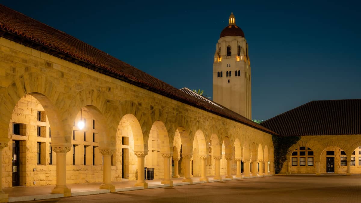 La icónica Torre Hoover de la Universidad Stanford iluminada de noche, elevándose sobre la arcada de piedra arenisca con arcos románicos del patio principal.