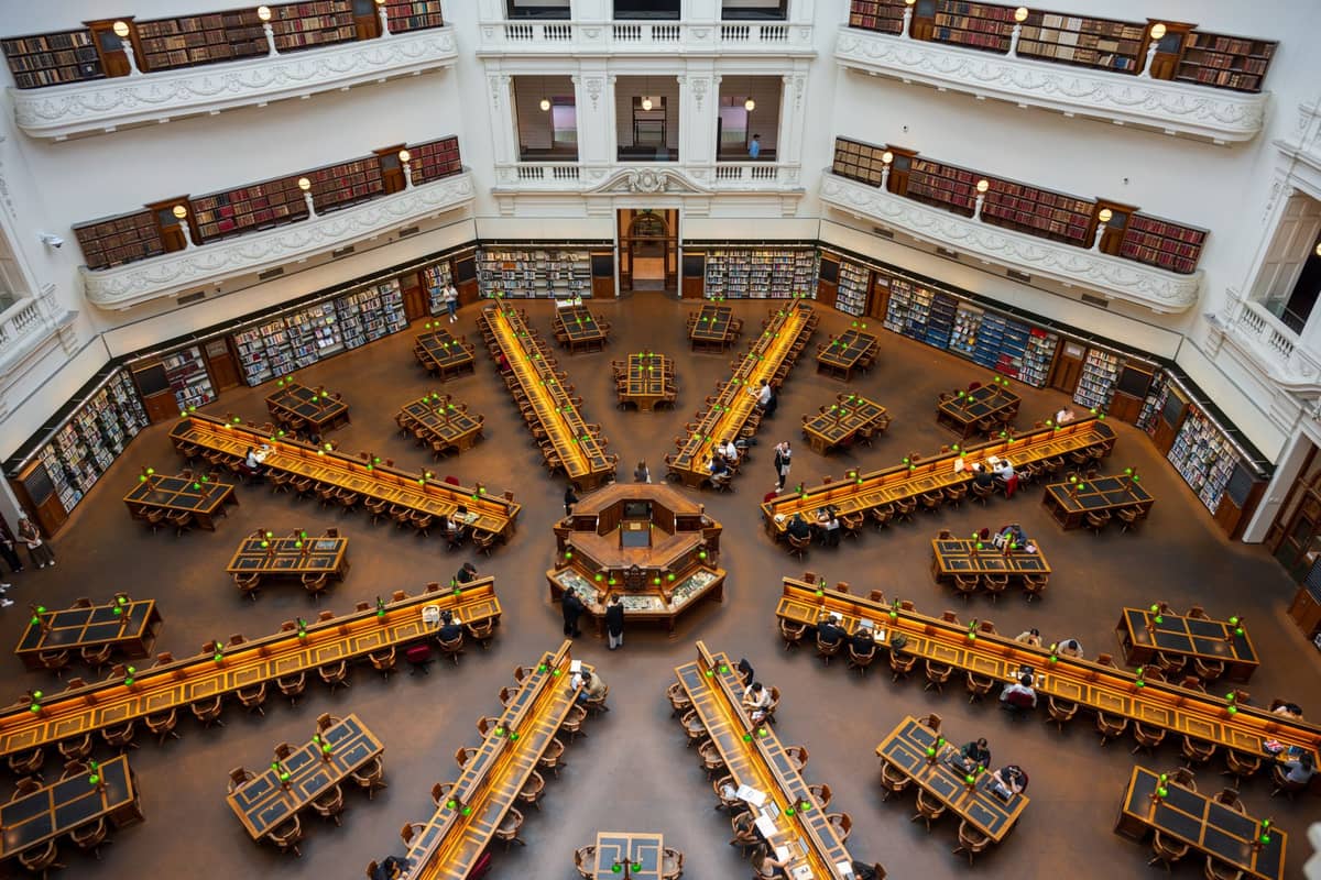 Vista aérea y panorámica de la icónica Sala de Lectura con cúpula de la State Library Victoria, con sus escritorios de madera oscura, lámparas de lectura verdes y balcones blancos repletos de estanterías.