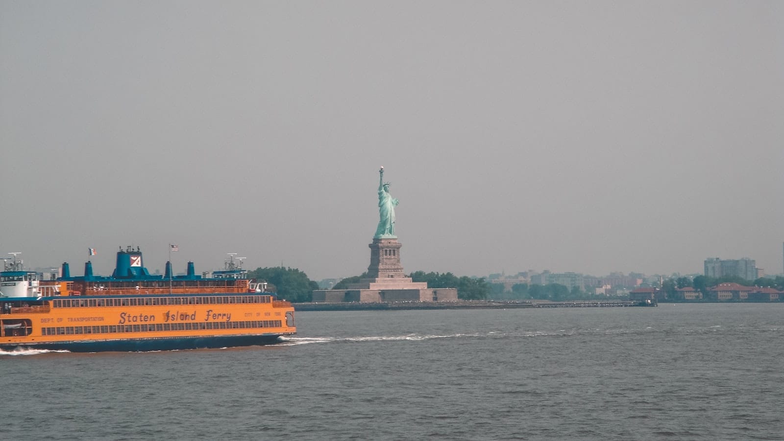 Un traghetto arancione dello Staten Island Ferry attraversa il porto di New York con la Statua della Libertà visibile in lontananza in una giornata velata.